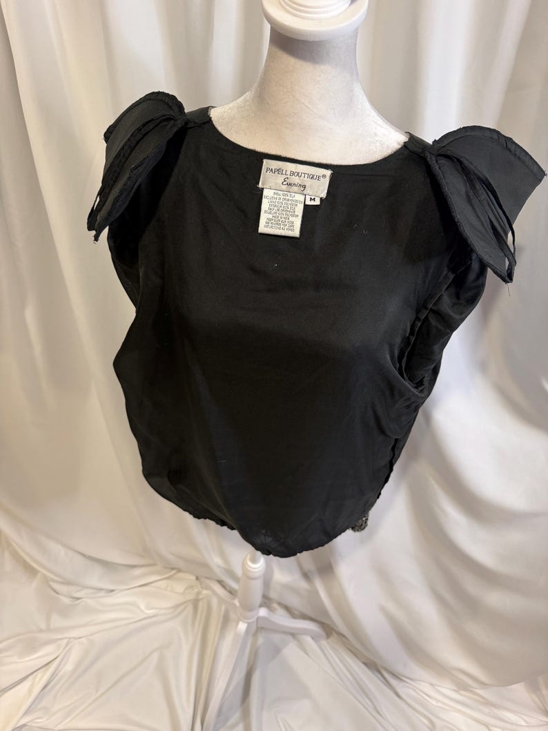 May include: A black sleeveless top with structured, pleated shoulder details and a round neckline. The interior label reads Papell Boutique Evening, size M. The garment features a solid black fabric construction with a gathered hemline at the waist.