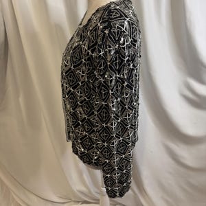 May include: A long-sleeved black jacket featuring an intricate geometric pattern created with silver beads and sequins. The garment has a round neckline and a straight hem, with the entire surface covered in a repeating diamond-shaped design.