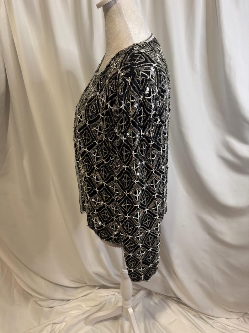 May include: A long-sleeved black jacket featuring an intricate geometric pattern created with silver beads and sequins. The garment has a round neckline and a straight hem, with the entire surface covered in a repeating diamond-shaped design.