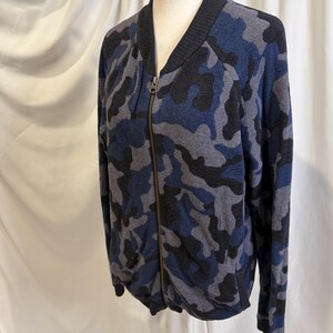 K&ouml;nnte beinhalten: Eine lang&auml;rmelige Bomberjacke mit Rei&szlig;verschluss in einem blauen, schwarzen und grauen Tarnmuster. Die Jacke hat einen schwarzen gerippten Kragen, B&uuml;ndchen und Saum sowie einen zentralen Metallrei&szlig;verschluss vom Halsausschnitt bis zum Saum.