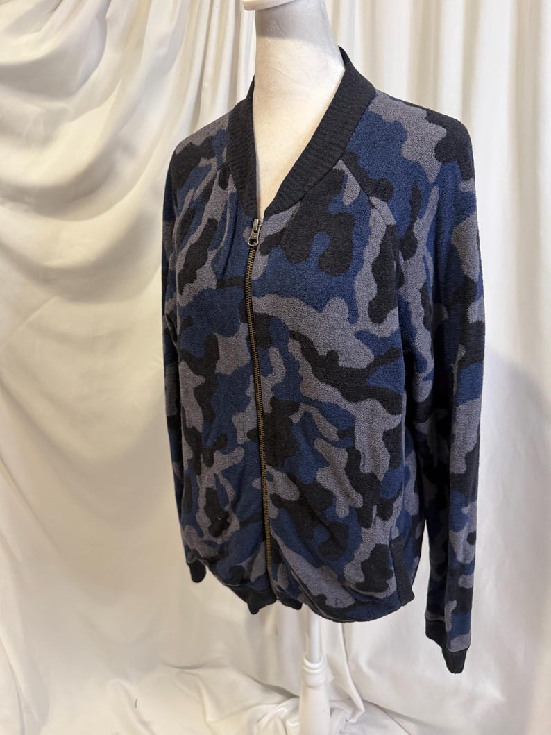 K&ouml;nnte beinhalten: Eine lang&auml;rmelige Bomberjacke mit Rei&szlig;verschluss in einem blauen, schwarzen und grauen Tarnmuster. Die Jacke hat einen schwarzen gerippten Kragen, B&uuml;ndchen und Saum sowie einen zentralen Metallrei&szlig;verschluss vom Halsausschnitt bis zum Saum.
