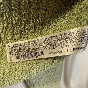 May include: Close-up of a light green, textured fabric. A white tag with black text is visible, including the brand name "INDITEX" and a barcode. The fabric appears soft and plush.