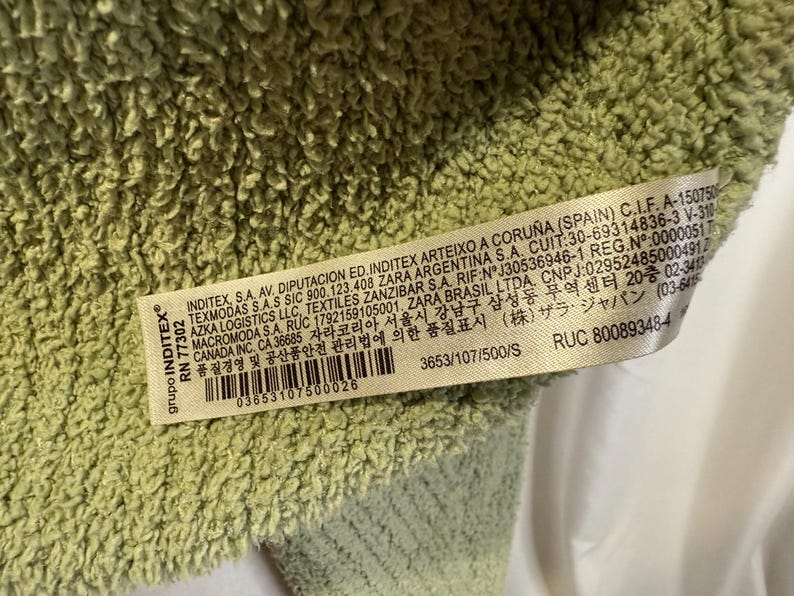 May include: Close-up of a light green, textured fabric. A white tag with black text is visible, including the brand name "INDITEX" and a barcode. The fabric appears soft and plush.