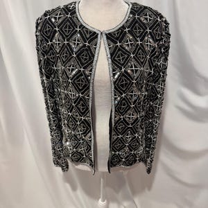 May include: A black open-front jacket featuring an intricate geometric pattern created with silver beads and sequins. The garment has long sleeves, a round neckline, and a structured fit, with the beadwork covering the entire front and sleeves.