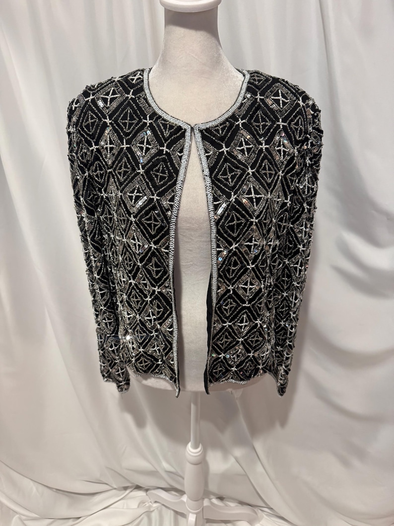 May include: A black open-front jacket featuring an intricate geometric pattern created with silver beads and sequins. The garment has long sleeves, a round neckline, and a structured fit, with the beadwork covering the entire front and sleeves.