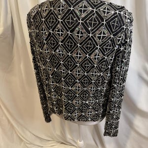 May include: A black long-sleeved jacket featuring an intricate geometric pattern created with silver beads, clear sequins, and white seed beads. The design consists of repeating diamond shapes outlined in white beads with silver bead accents.