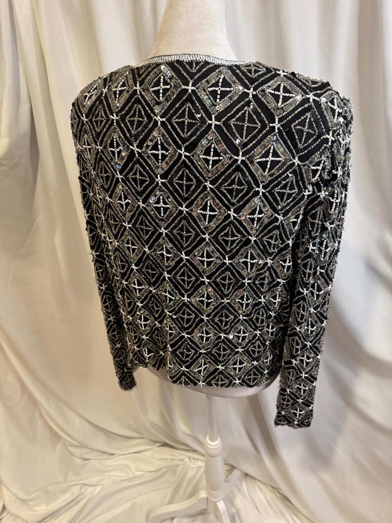 May include: A black long-sleeved jacket featuring an intricate geometric pattern created with silver beads, clear sequins, and white seed beads. The design consists of repeating diamond shapes outlined in white beads with silver bead accents.