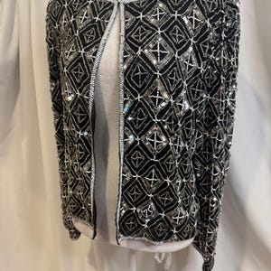 May include: A black long-sleeve jacket featuring an intricate geometric pattern created with silver sequins and white beads. The garment has a round neckline, an open front design, and is fully embellished with a repeating diamond motif throughout.