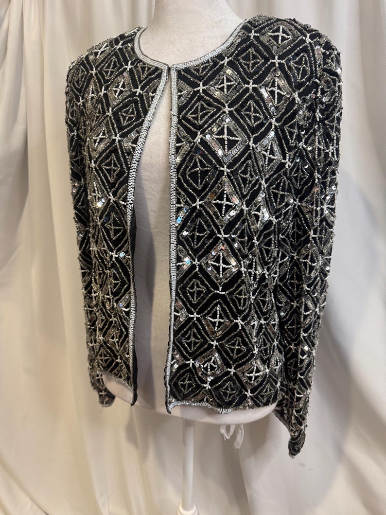 May include: A black long-sleeve jacket featuring an intricate geometric pattern created with silver sequins and white beads. The garment has a round neckline, an open front design, and is fully embellished with a repeating diamond motif throughout.
