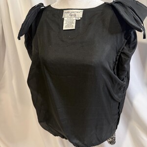 May include: A black sleeveless top with structured, pleated shoulders and a round neckline. The interior features a white label with the text Papell Boutique Evening and size M. The garment has a gathered, elasticized hemline at the waist.