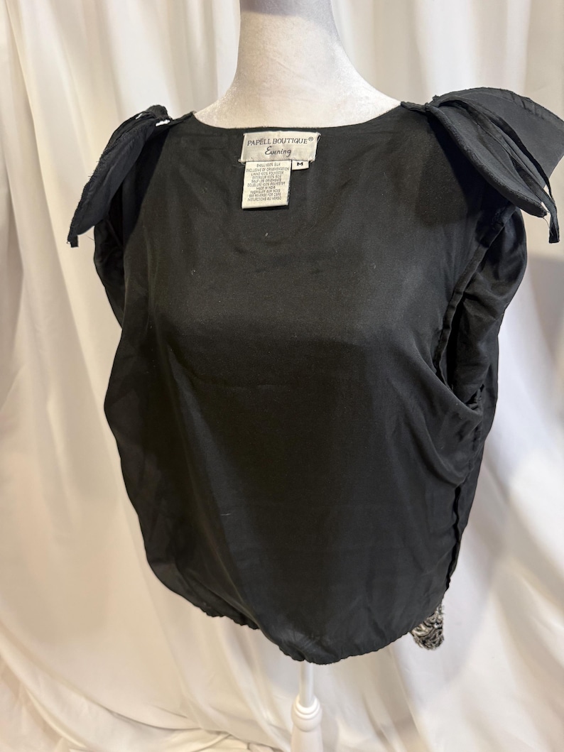 May include: A black sleeveless top with structured, pleated shoulders and a round neckline. The interior features a white label with the text Papell Boutique Evening and size M. The garment has a gathered, elasticized hemline at the waist.