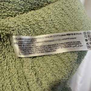 May include: Close-up of a light green, textured fabric. A white tag is attached, displaying text in multiple languages, including English, Spanish, and Japanese, detailing the fabric's composition and origin.