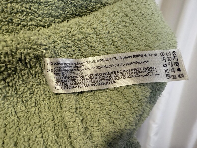 May include: Close-up of a light green, textured fabric. A white tag is attached, displaying text in multiple languages, including English, Spanish, and Japanese, detailing the fabric's composition and origin.