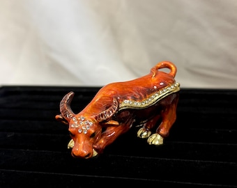May include: A decorative orange and gold ox figurine with rhinestone accents. The ox is in a crouching position, with its horns and hooves detailed in gold. The figurine is set against a black background.