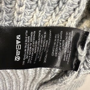 Puede incluir: Primer plano de una etiqueta de ropa negra con texto blanco y s&iacute;mbolos de cuidado, sobre una tela de punto gris claro. La etiqueta proporciona instrucciones de lavado, incluyendo lavar a mano en fr&iacute;o y secar en plano.