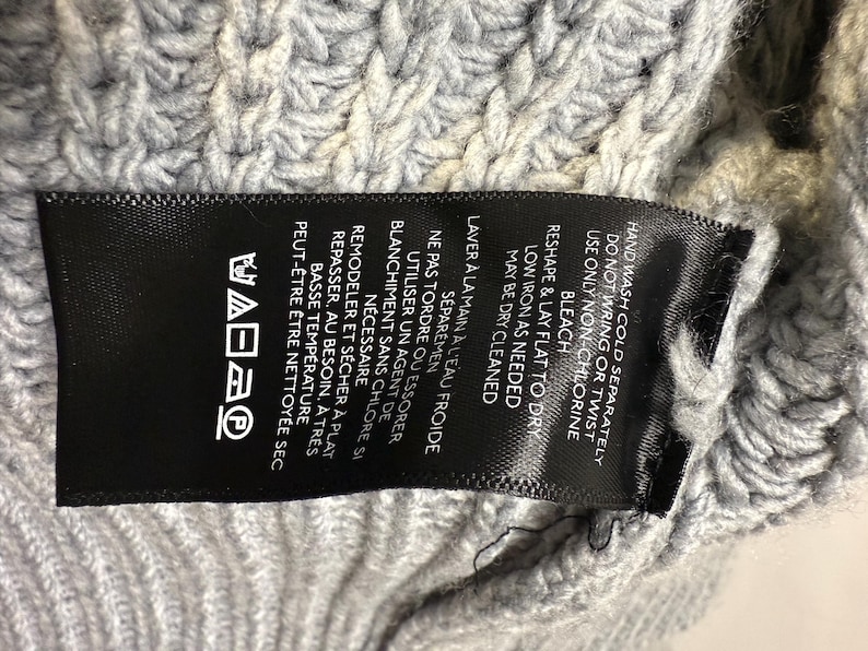 Puede incluir: Primer plano de una etiqueta de ropa negra con texto blanco y s&iacute;mbolos de cuidado, sobre una tela de punto gris claro. La etiqueta proporciona instrucciones de lavado, incluyendo lavar a mano en fr&iacute;o y secar en plano.