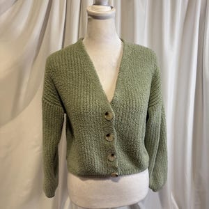 May include: A sage green cardigan sweater with a V-neckline and button closure. The sweater is displayed on a mannequin against a white curtain backdrop. The cardigan has a textured knit design.