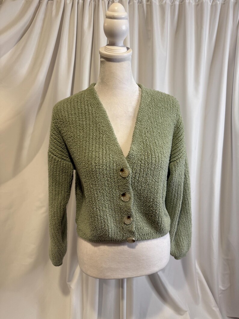 May include: A sage green cardigan sweater with a V-neckline and button closure. The sweater is displayed on a mannequin against a white curtain backdrop. The cardigan has a textured knit design.