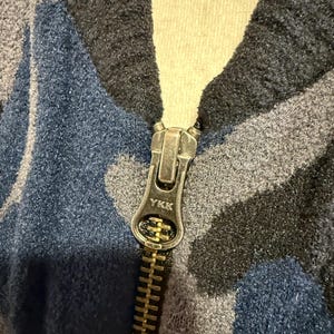 K&ouml;nnte beinhalten: Eine Fleecejacke mit einem blauen, grauen und schwarzen Tarnmuster. Das Kleidungsst&uuml;ck verf&uuml;gt &uuml;ber einen metallischen Rei&szlig;verschluss mit einem silberfarbenen Schiebergriff, auf dem das YKK-Logo eingepr&auml;gt ist, mittig am Kragen.