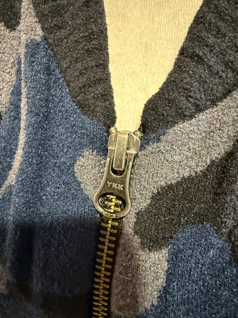 K&ouml;nnte beinhalten: Eine Fleecejacke mit einem blauen, grauen und schwarzen Tarnmuster. Das Kleidungsst&uuml;ck verf&uuml;gt &uuml;ber einen metallischen Rei&szlig;verschluss mit einem silberfarbenen Schiebergriff, auf dem das YKK-Logo eingepr&auml;gt ist, mittig am Kragen.