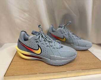 May include: A pair of light blue athletic shoes with a yellow and red swoosh logo. The shoes have a yellow sole and are displayed on a wooden surface. The shoes have the text "GT-D" on the tongue.
