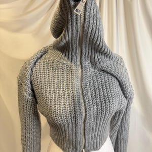Puede incluir: Un su&eacute;ter corto gris con capucha y cremallera completa. El su&eacute;ter tiene mangas largas y una textura de punto acanalado. La cremallera es plateada. El su&eacute;ter est&aacute; sostenido por una mano enguantada.