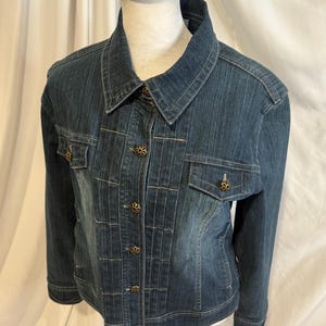 May include: A dark blue denim jacket with a classic collar and button-up front. The jacket features two chest pockets and decorative buttons. The jacket is displayed on a mannequin.