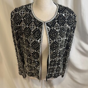 May include: A black open-front jacket featuring an intricate geometric pattern created with silver sequins and beads. The garment has long sleeves, a round neckline, and a repeating diamond motif across the entire surface, finished with silver trim.