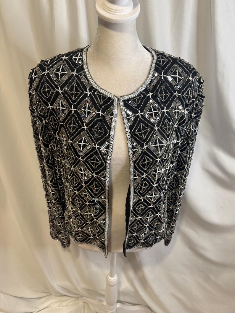 May include: A black open-front jacket featuring an intricate geometric pattern created with silver sequins and beads. The garment has long sleeves, a round neckline, and a repeating diamond motif across the entire surface, finished with silver trim.