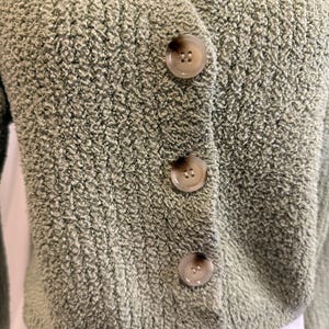 May include: A sage green cardigan with a V-neck and three large, round buttons down the front. The sweater has a textured, fuzzy knit fabric. The buttons are a light brown color. The cardigan is cropped and appears to be made of a soft material.