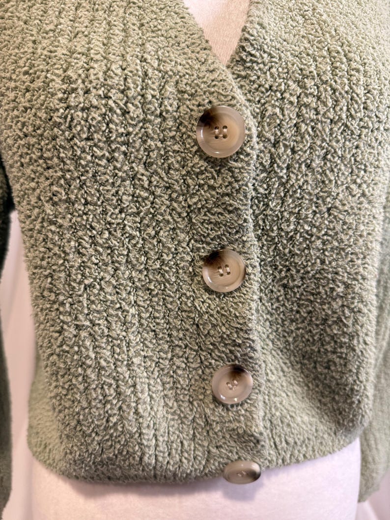 May include: A sage green cardigan with a V-neck and three large, round buttons down the front. The sweater has a textured, fuzzy knit fabric. The buttons are a light brown color. The cardigan is cropped and appears to be made of a soft material.