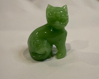 May include: A small, green glass cat figurine in a seated position. The cat has detailed features, including ears, a face, and a curled tail. The figurine is a vibrant shade of green and sits on a white surface.