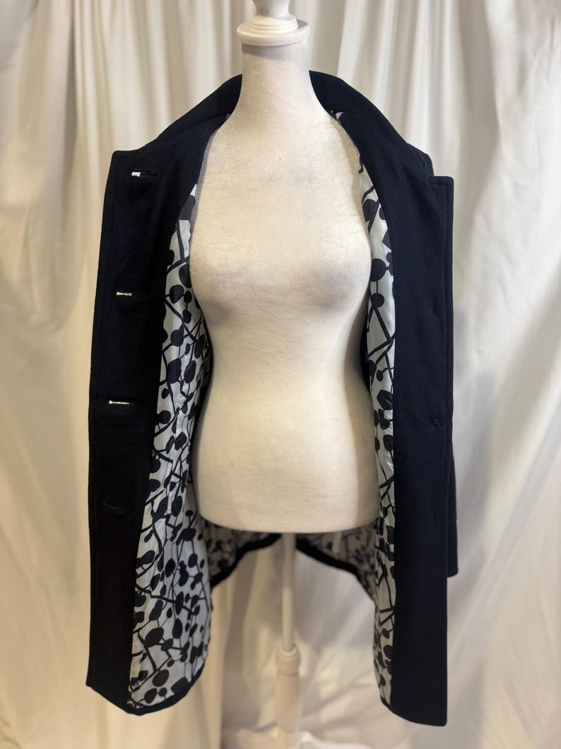 Puede incluir: Un abrigo azul marino con forro estampado en blanco y negro. El abrigo est&aacute; abotonado y se muestra en un maniqu&iacute;. El forro presenta un dise&ntilde;o abstracto repetitivo. El exterior del abrigo es de color oscuro liso.