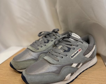 May include: A pair of gray Reebok sneakers with white accents and a navy blue sole. The sneakers are on a light wood surface. The Reebok logo is visible on the side of the shoe.