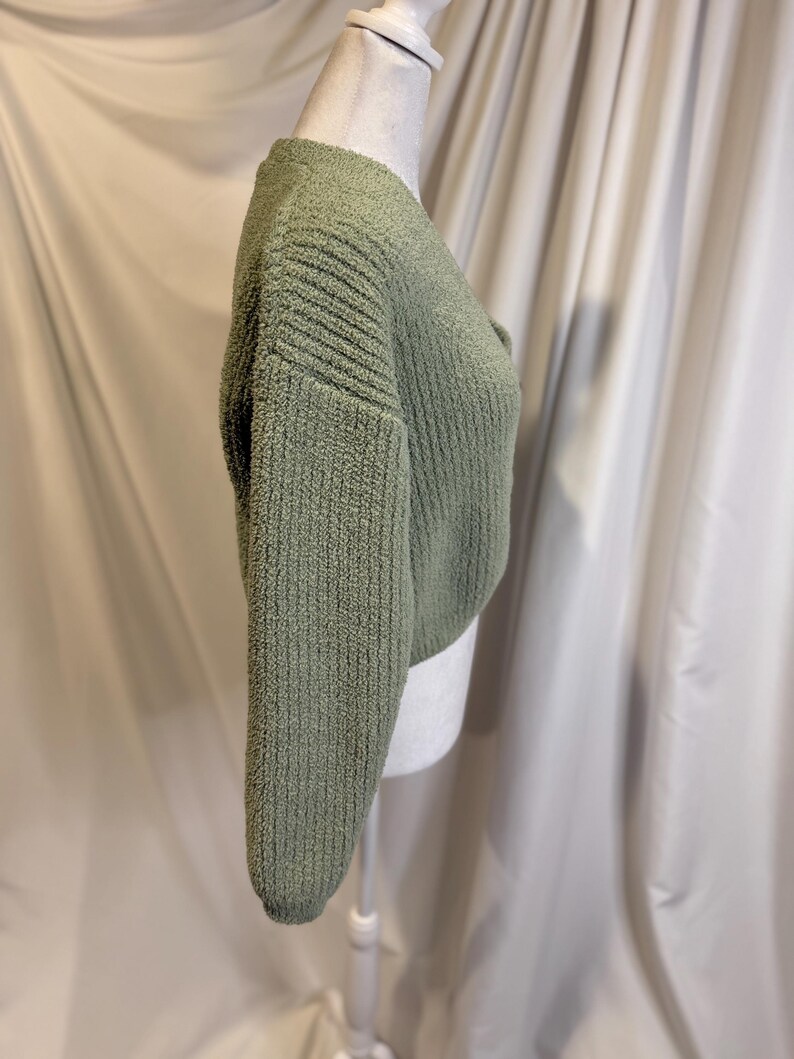May include: A sage green, cropped sweater with a textured, ribbed knit design. The long sleeves and the body of the sweater have a soft, fuzzy appearance. The sweater is displayed on a mannequin.