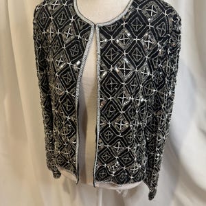 May include: A black long-sleeve jacket featuring an intricate geometric pattern created with silver and white beads and sequins. The garment has a round neckline, a straight open front, and is fully embellished with a repeating diamond motif.