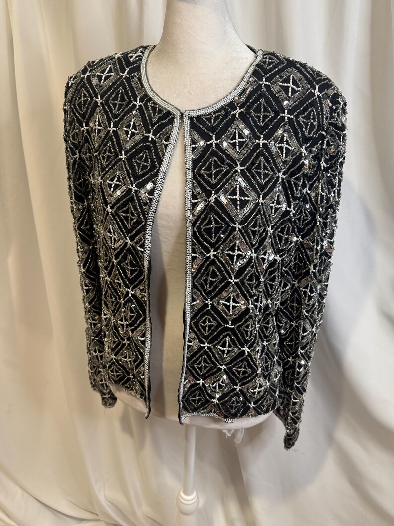 May include: A black long-sleeve jacket featuring an intricate geometric pattern created with silver and white beads and sequins. The garment has a round neckline, a straight open front, and is fully embellished with a repeating diamond motif.