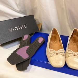 May include: A pair of beige ballet flats with bow details, alongside a pair of insoles and a black box with the brand name "VIONIC" in white. The shoes are on a blue box.