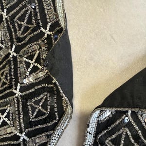 May include: A black garment featuring intricate geometric patterns created with silver sequins, black sequins, and small white beads. The design consists of repeating square motifs with cross-hatch detailing, finished with a white beaded border along the edge.
