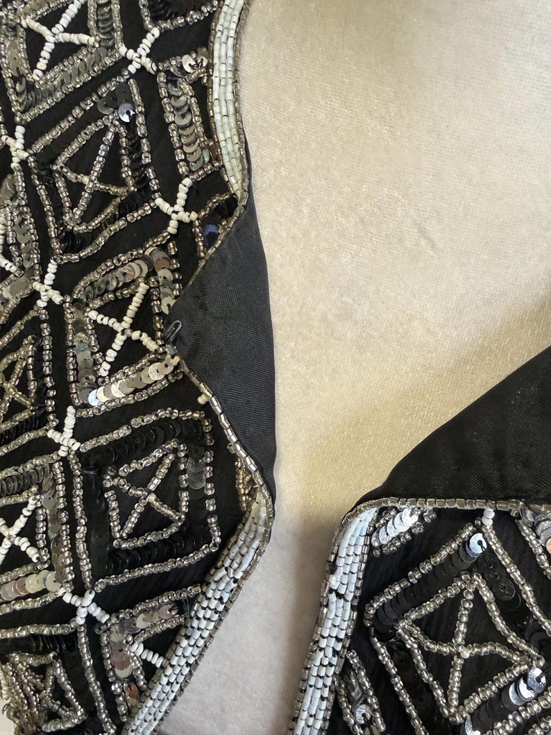 May include: A black garment featuring intricate geometric patterns created with silver sequins, black sequins, and small white beads. The design consists of repeating square motifs with cross-hatch detailing, finished with a white beaded border along the edge.
