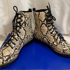 May include: A pair of ankle-high lace-up boots featuring a beige and black snakeskin pattern. The boots have black laces, a side zipper closure, a rounded toe, and a thick black lug sole with visible stitching along the edge of the base.