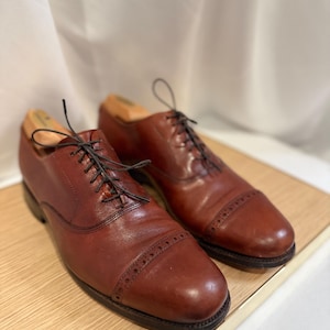 May include: A pair of brown leather dress shoes with black laces and a cap-toe design. The shoes are displayed on a light-colored wooden surface, showcasing their classic style and craftsmanship. Shoe trees are inserted in the shoes.