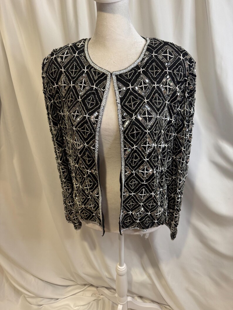 May include: A black open-front jacket featuring an intricate geometric pattern created with silver and white beads and sequins. The garment has long sleeves, a round neckline, and a repeating diamond motif covering the entire surface.