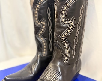 May include: A pair of black leather cowboy boots featuring a pointed toe, a stacked heel, and decorative white stitching patterns along the shaft and vamp. The boots have a tall silhouette with pull tabs at the top and a smooth, polished finish.