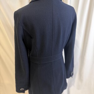 Puede incluir: Un abrigo azul marino con cuello con solapas y cintur&oacute;n en la cintura. El abrigo tiene mangas largas y dos botones en cada pu&ntilde;o. La parte trasera del abrigo tiene un dise&ntilde;o a medida con costuras verticales.