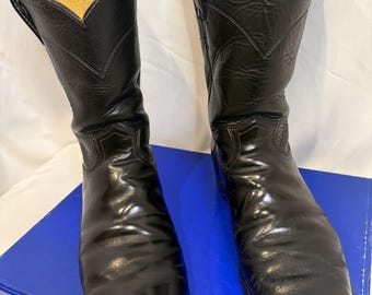 May include: A pair of black leather cowboy boots featuring a classic mid-calf height, pointed toes, and a distinct V-shaped collar design. The boots have a smooth, polished finish with visible stitching details along the shaft and foot area.