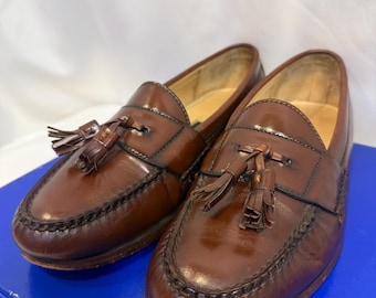 May include: A pair of brown leather tassel loafers featuring a moccasin-style stitched toe, a polished finish, and decorative leather tassels on the vamp. The interior lining is light tan, and the shoes are displayed on a solid blue surface.