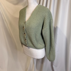 May include: A sage green cardigan with a V-neck and long sleeves. The cropped sweater has a textured knit and is fastened with four round, light brown buttons. The garment is displayed on a white mannequin.