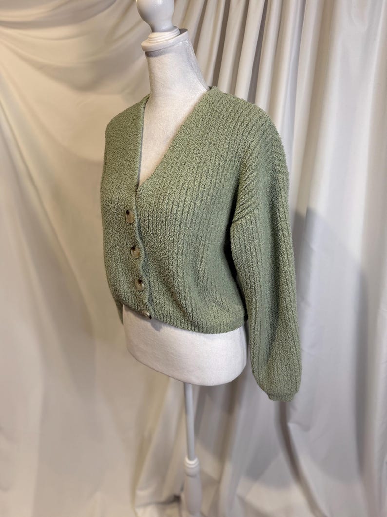 May include: A sage green cardigan with a V-neck and long sleeves. The cropped sweater has a textured knit and is fastened with four round, light brown buttons. The garment is displayed on a white mannequin.