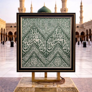 May include: A framed piece of artwork with white Arabic calligraphy on a green background. The frame is displayed on a gold stand with a marble base, and prayer beads are on the base. The background shows a mosque.